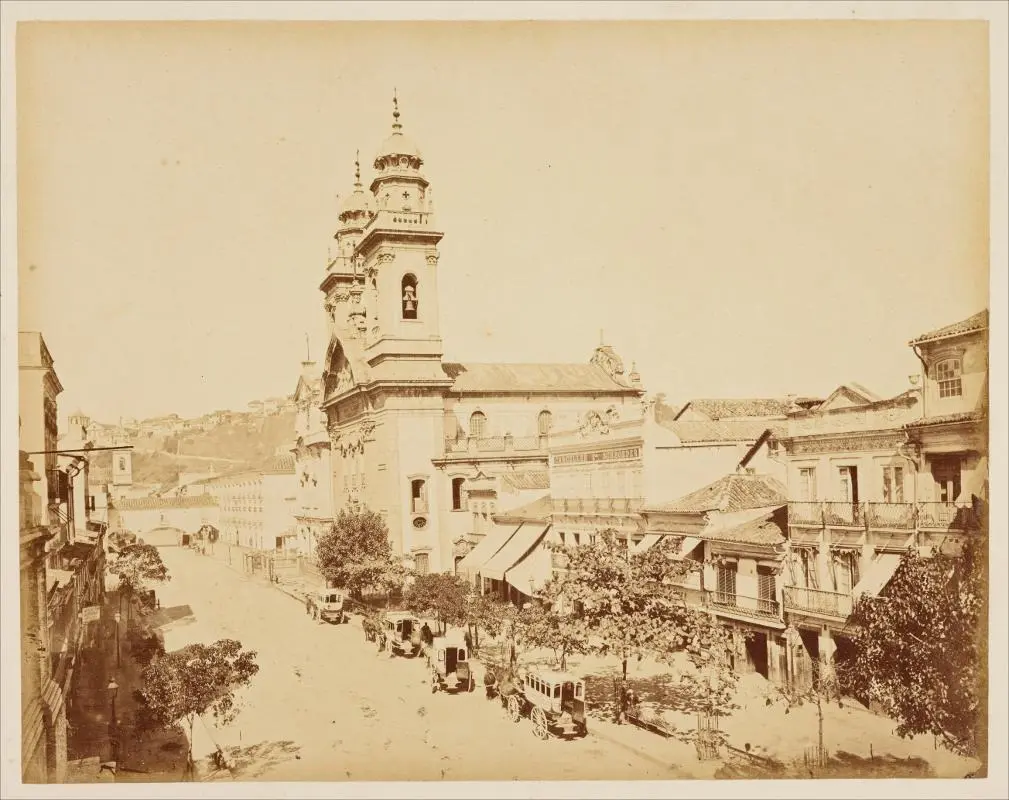 Uma praça urbana histórica com edifícios, igreja e árvores.