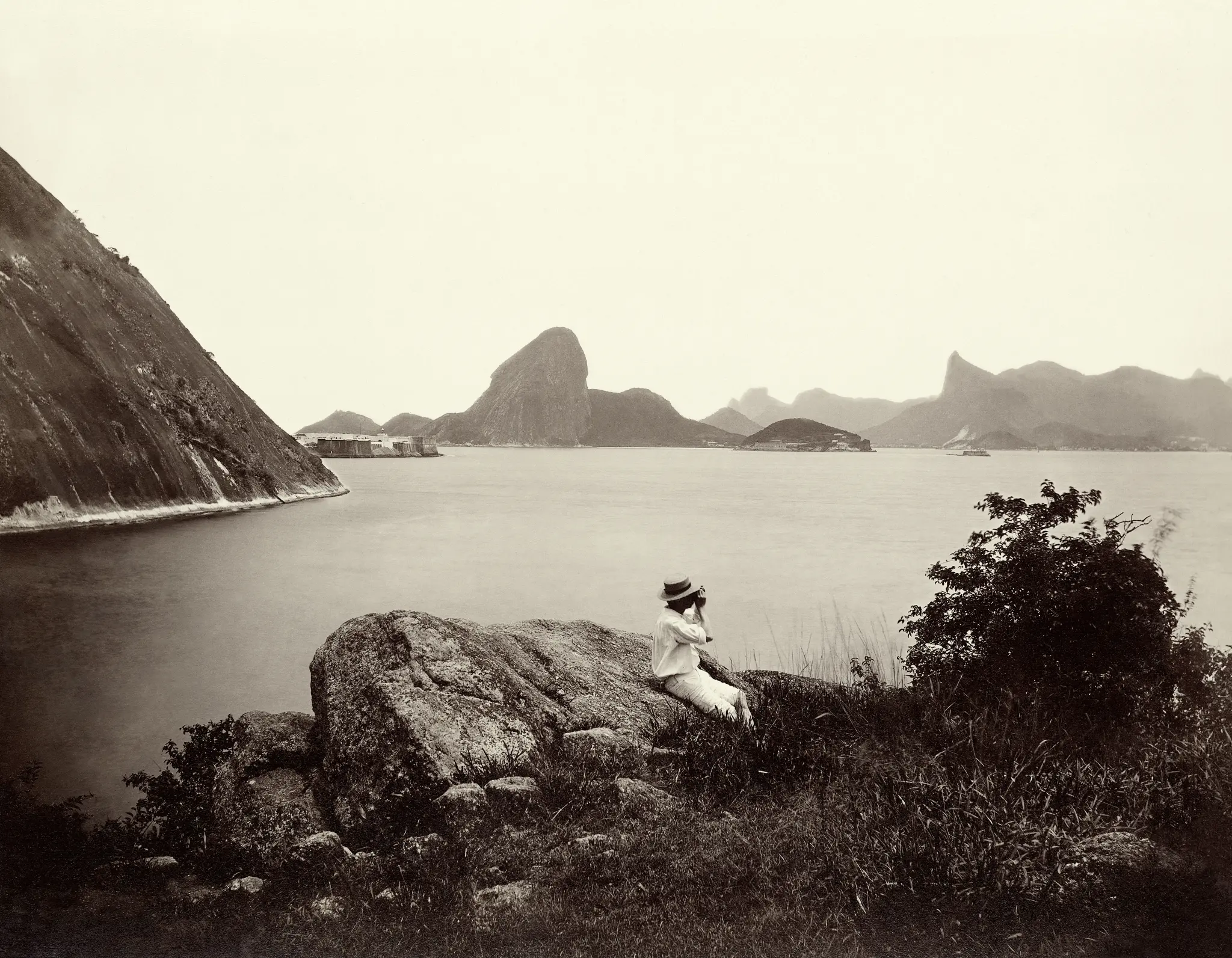 Paisagem costeira antiga com rocha íngreme, ilhas, montanhas, homem sentado, água calma e vegetação densa.