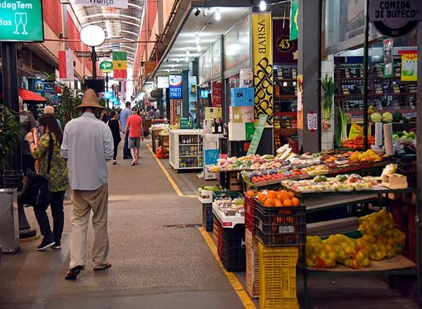 Mercado local com pessoas caminhando, produtos frescos e refrigerador de bebidas.