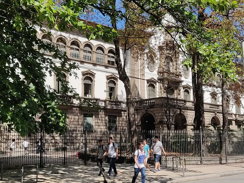 Prédio histórico em frente à árvore, com cerca metálica e pessoas caminhando.