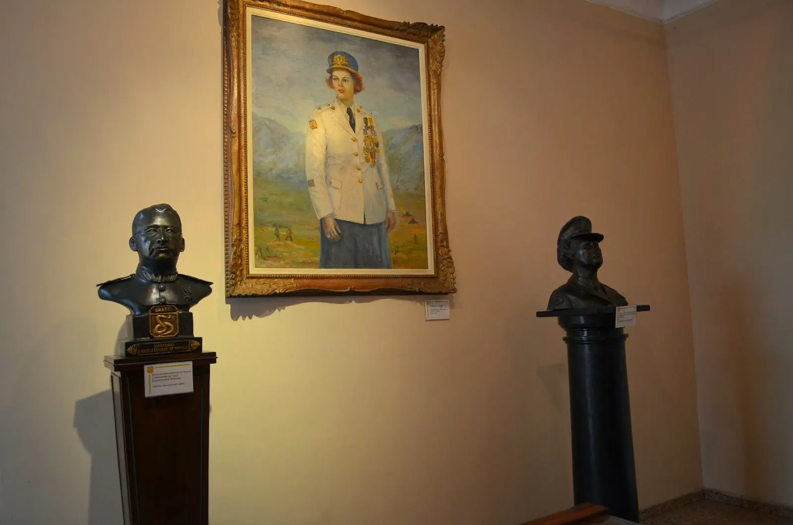 Busto feminino militar e dois bustos em pedestais diante de uma pintura.