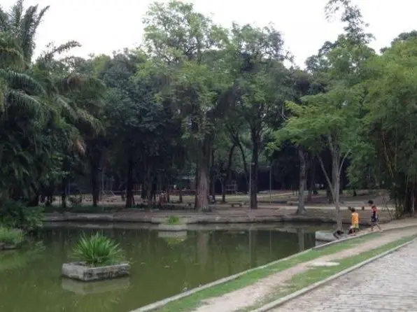 Parque com árvores verdes, lago calmo e pessoas caminhando.