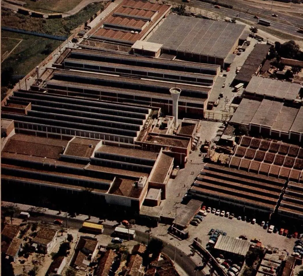 Complexo industrial com edifícios em linhas paralelas e torre cilíndrica branca.
