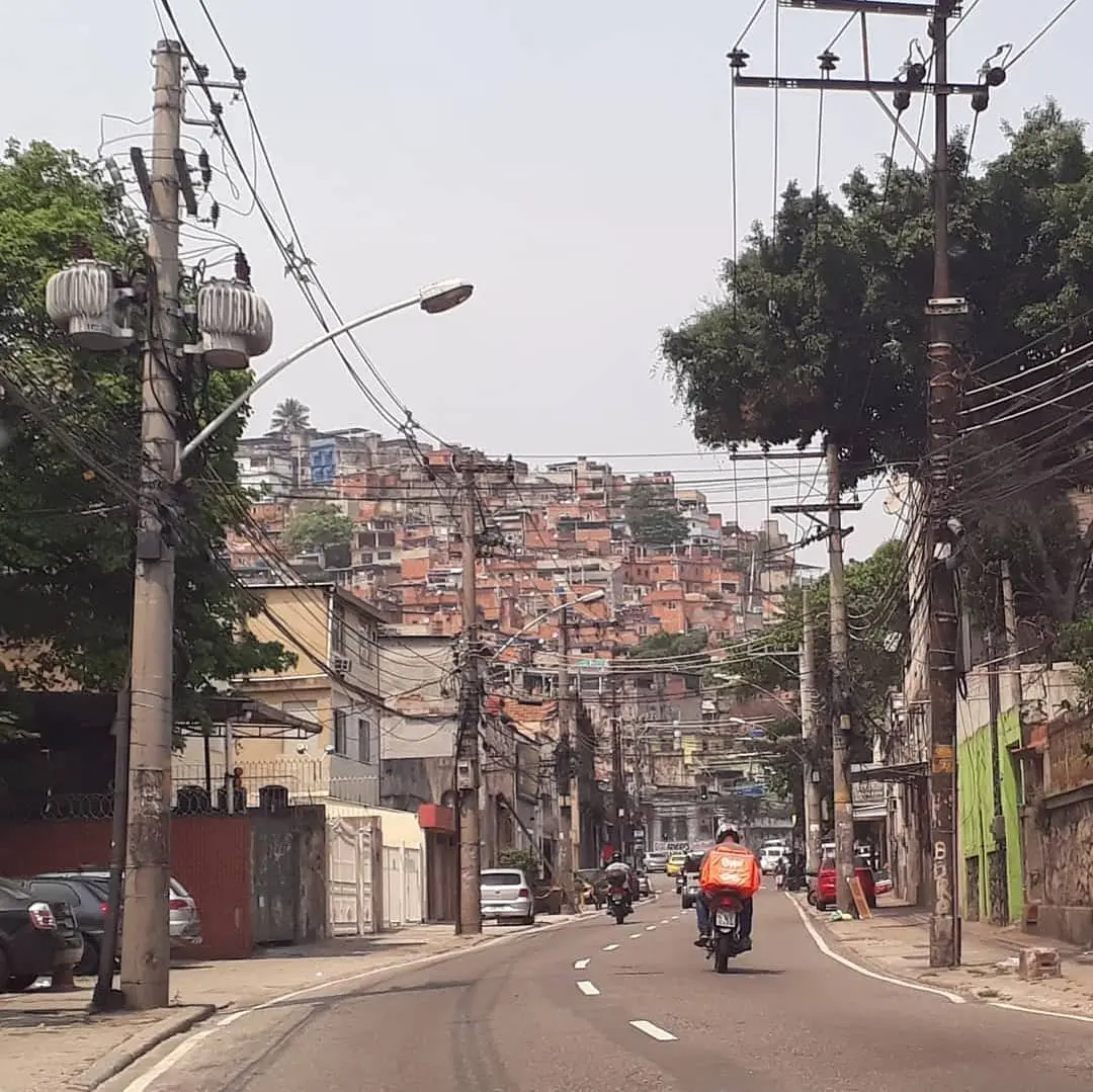 Uma rua urbana inclinada com motociclistas, casas coloridas e árvores.
