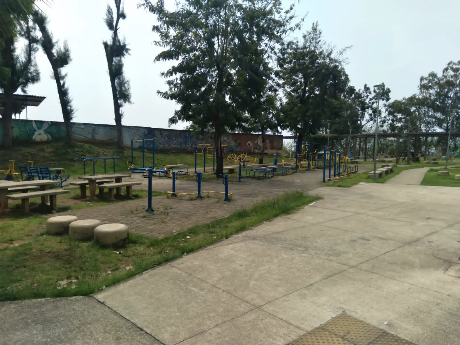 Parque com mesas, bancos, equipamentos de ginástica e árvores.