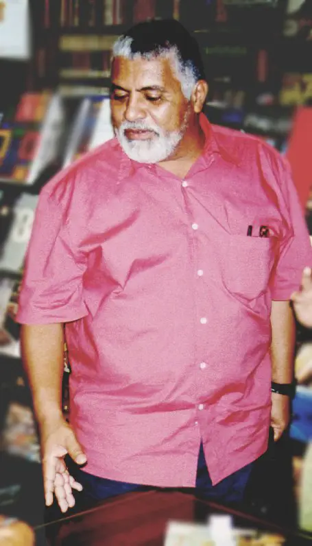 Homem com camisa rosa e barba branca em frente a estante de livros variados.