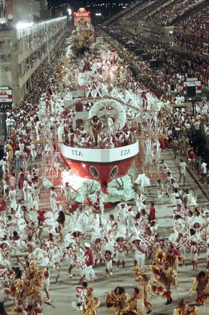 Barco flutuante "ITA" em desfile de Carnaval, multidões branco-vermelho dançando.