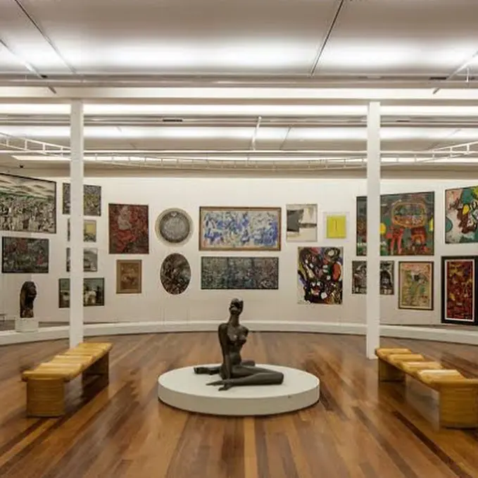 Sala de museu com obras de arte variadas e bancos de madeira.