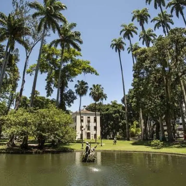 Parque com árvores e lago, edifício clássico, céu azul claro.