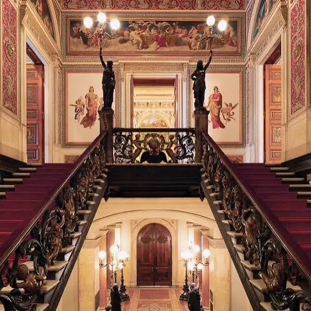 Interior de museu com escadaria, colunas femininas, painéis ornamentais e porta iluminada.