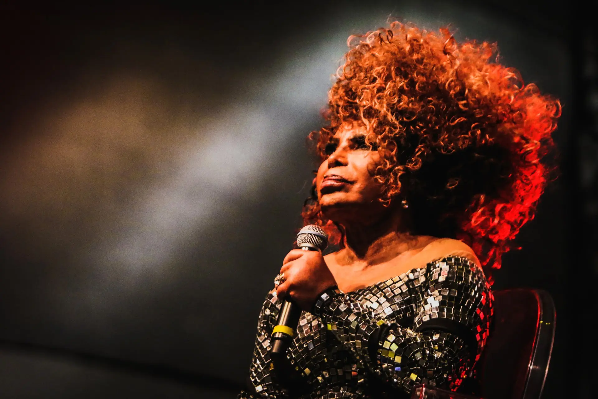 Mulher cantando ao vivo com vestido preto e microfone, fundo escuro com luz branca.