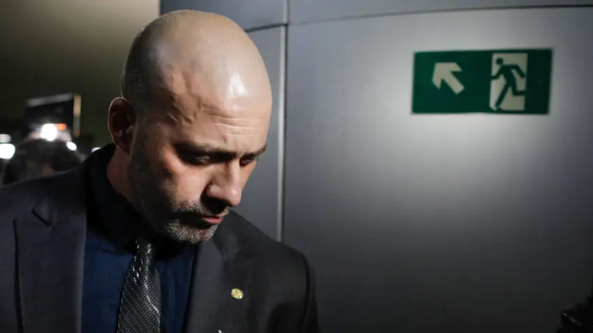 Homem de perfil em terno preto, olhando para baixo.