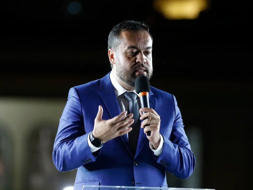 Homem falando em microfone, vestido com terno azul e camisa branca, fundo escuro com luz amarela.
