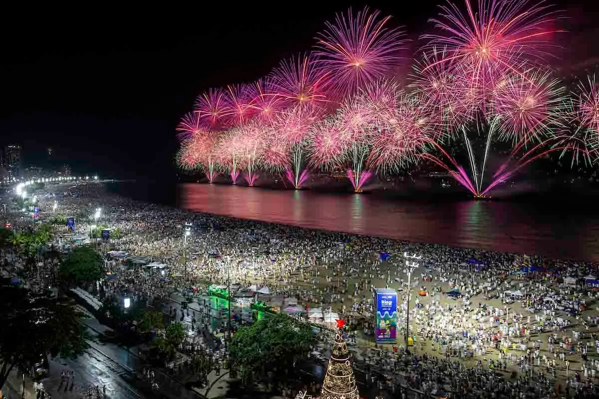 Multidão animada na praia de Copacabana durante Réveillon, com reflexo de fogos coloridos no rio.