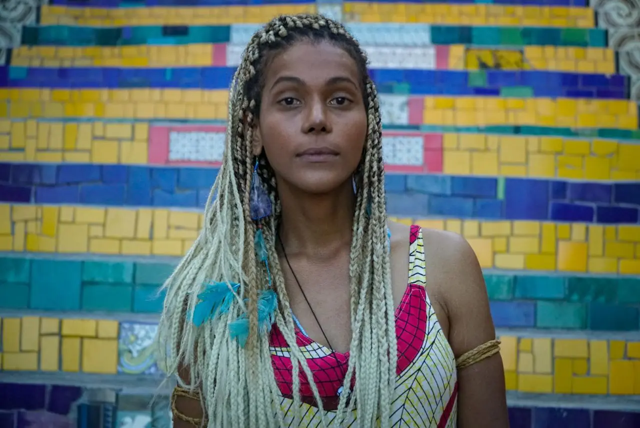 Uma mulher com tranças coloridas e top vibrante em frente a parede de azulejos amarelos, verdes e azuis.