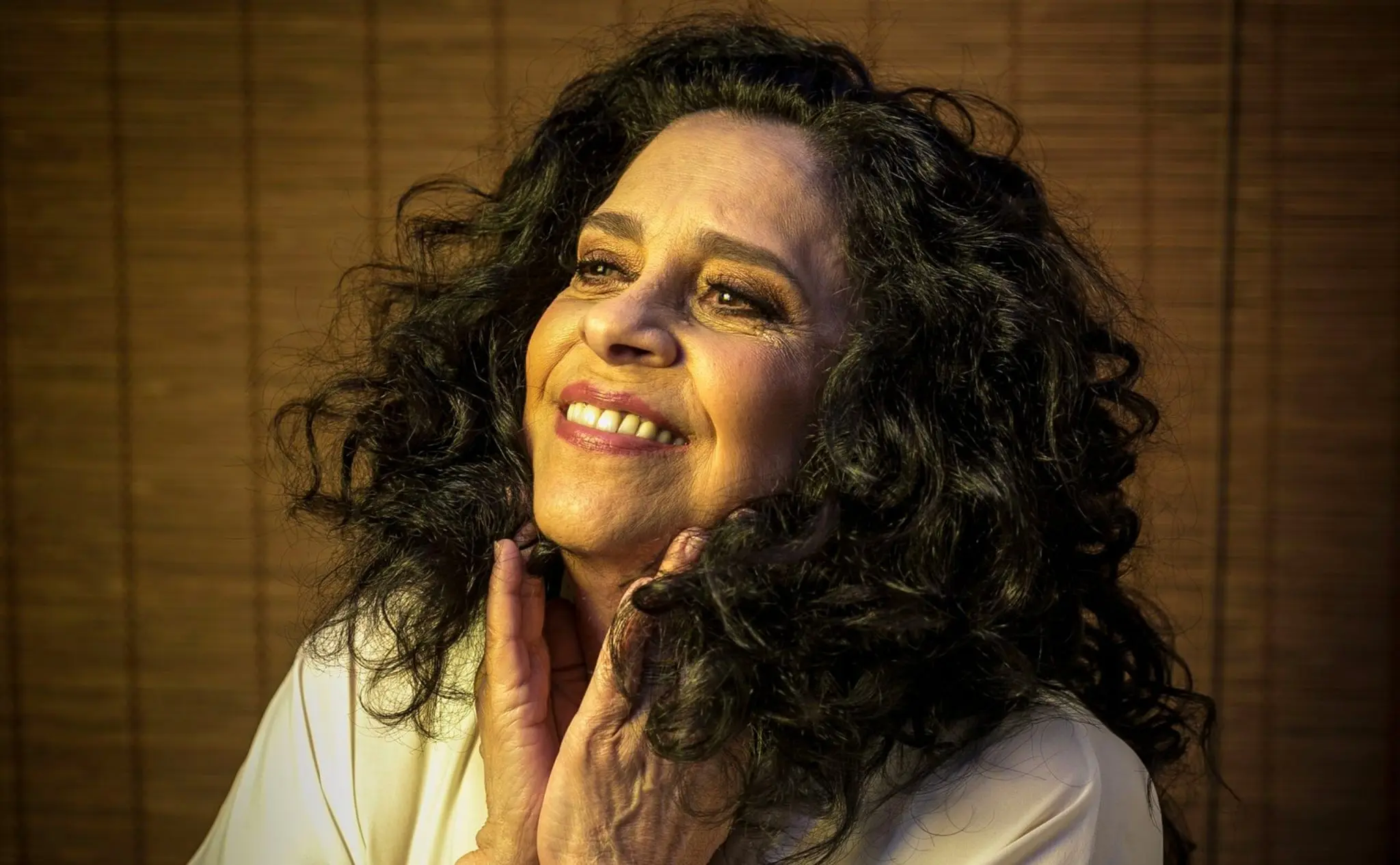 Uma mulher sorridente com cabelos cacheados pretos e blusa branca, atrás cortinas marrom escuro.