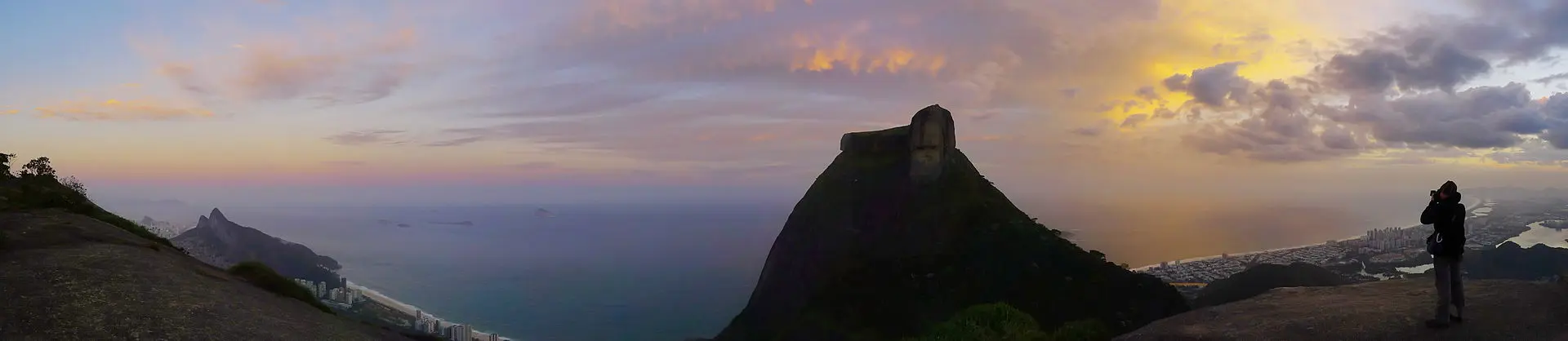 Paisagem costeira com montanha, cidade, rochedo e pessoa fotografando ao pôr-do-sol.