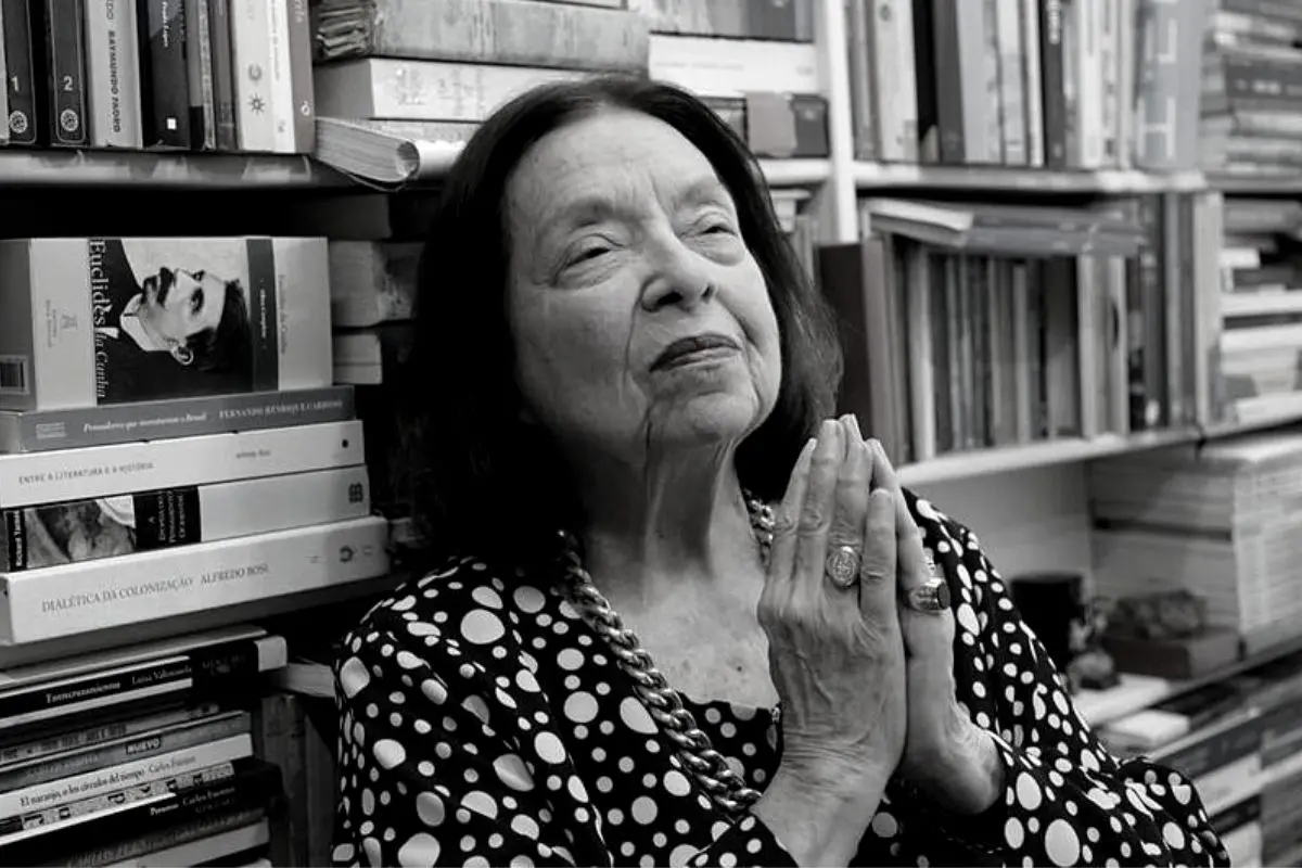Mulher em preto e branco, sentada, olhos fechados, mãos juntas em oração, blusa com pontos grandes e corrente de joia.