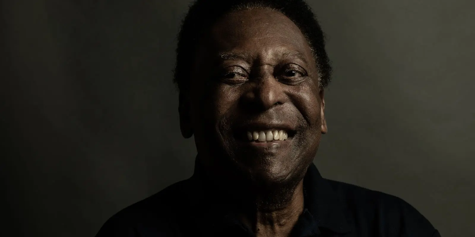 Homem sorrindo amplamente, camisa preta, fundo escuro.