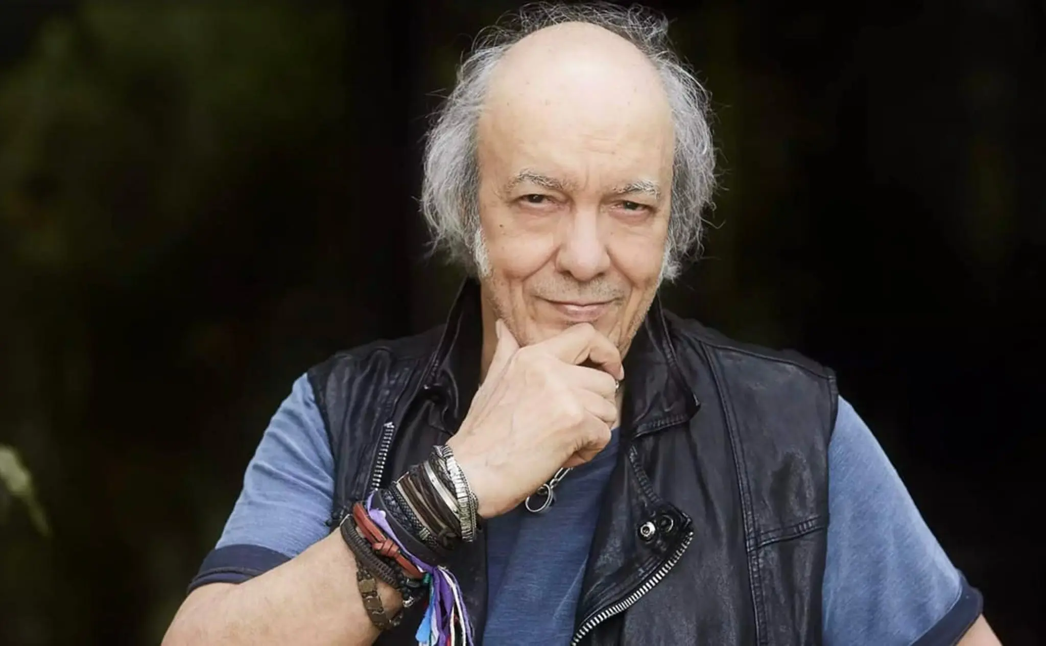 Homem sorrindo, camisa azul, jaqueta de couro preta, fundo verde e preto.