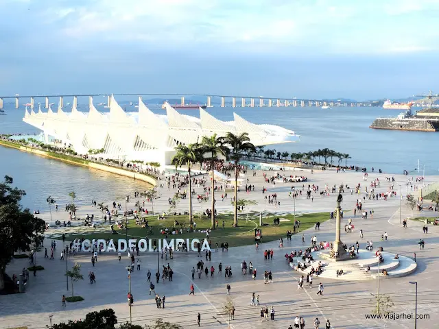 Edifício moderno com triângulos brancos, praça com pessoas caminhando, estátua centralizada, mar e ponte ao fundo, céu azul claro.