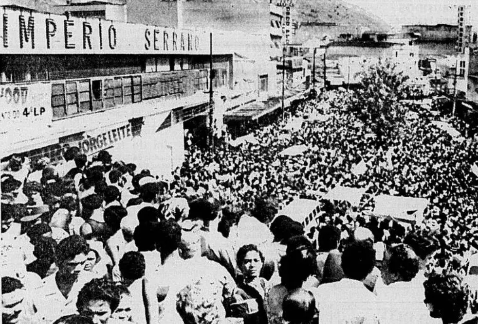 Multidão em rua urbana, edifício "Imperio Serrano", placa "007" e "Jorge Leite".