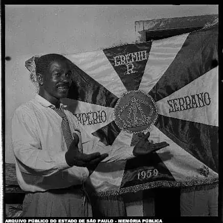 Homem sorridente com troféu, bandeira do Império Serrano de 1959 ao fundo.