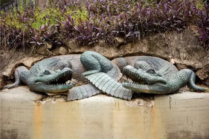 Duas esculturas de crocodilos em um muro com plantas roxas.