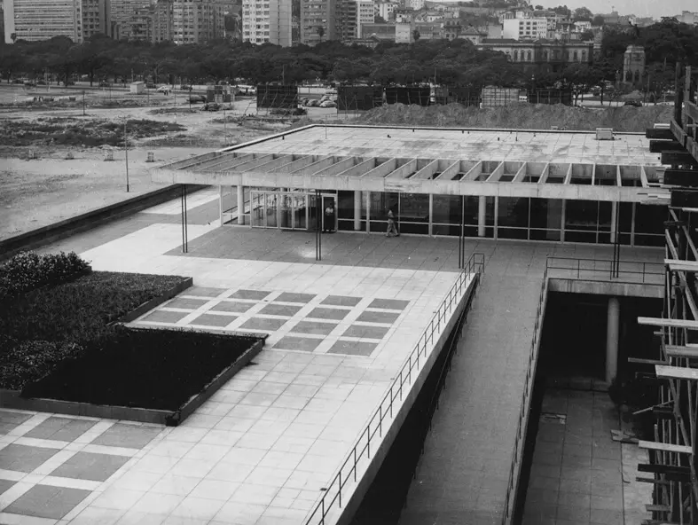 Edifício moderno com planta e área pavimentada à esquerda, fachada de vidro e estrutura metálica no centro, e escada externa com grades laterais à direita. Fundo com cidade e árvores.