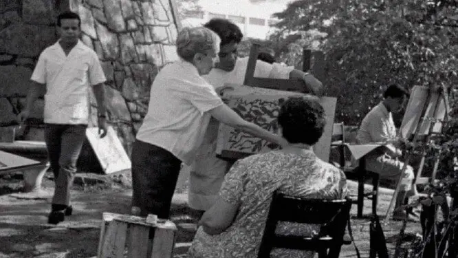 Homem caminhando com saco, mulher segurando quadro, dois homens sentados atrás, outro concentrado em trabalho, tudo em ambiente ao ar livre cercado por pedras e árvores.