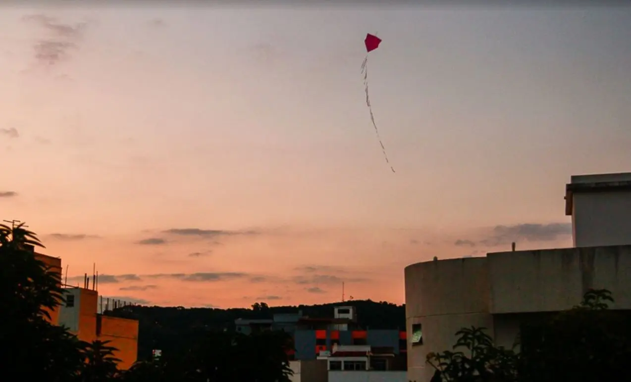Céu de pôr-do-sol com árvores, prédios e pipa vermelha.