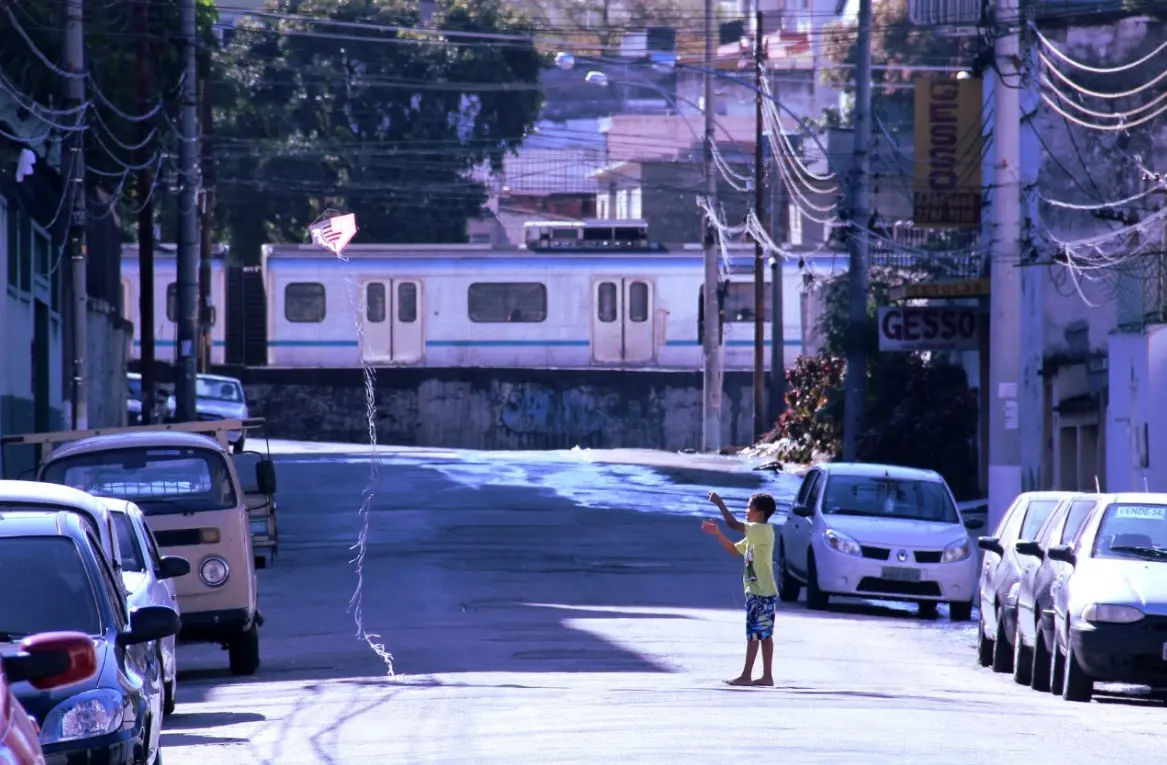 Menino na calçada olhando para trás, carro antigo à esquerda, trem abandonado ao fundo, placa "GESSO" à direita.