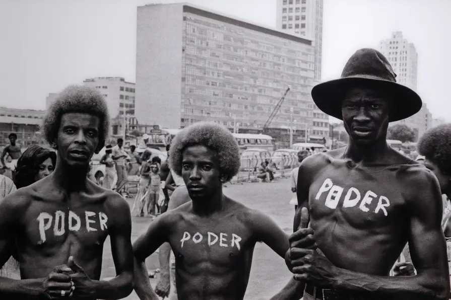 Três homens adultos nus do peito para baixo em preto e branco, com "PODER" escrito no tórax, em ambiente urbano.
