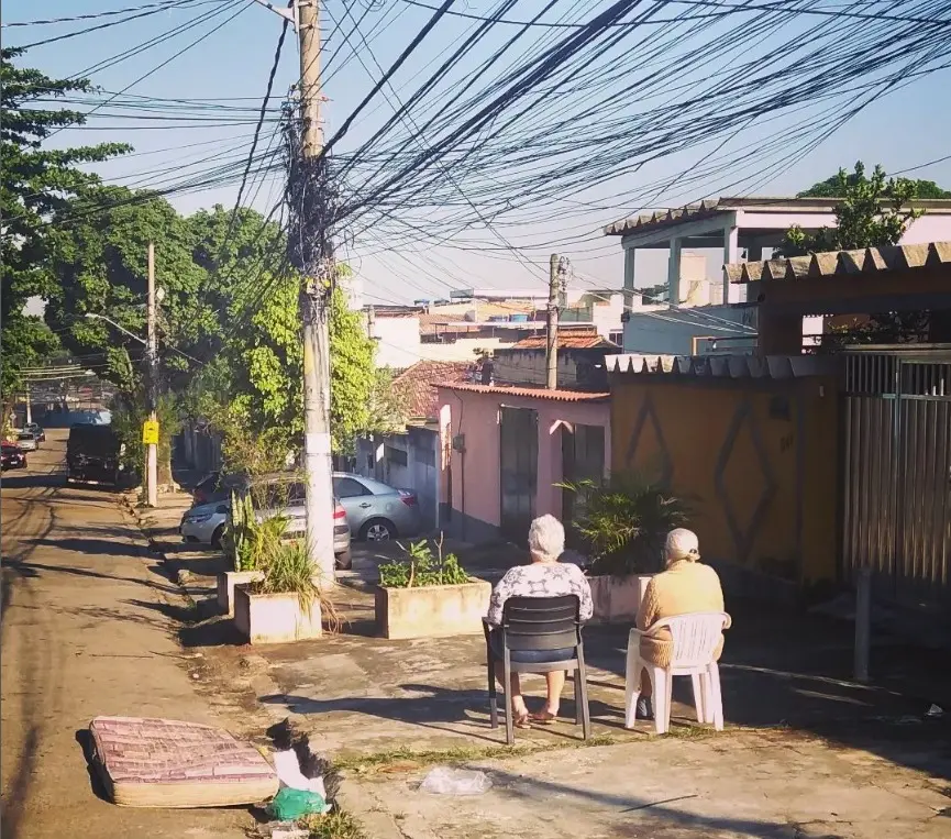 Rua em subúrbio com idosos sentados, árvores e casas coloridas.