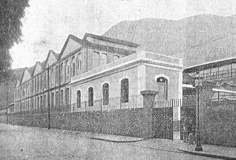 Prédio industrial antigo com cerca metálica e montanha ao fundo.