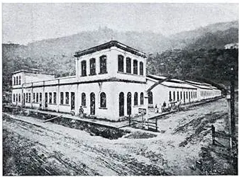 Edifício industrial com janelas horizontais, pátio e colina verde.