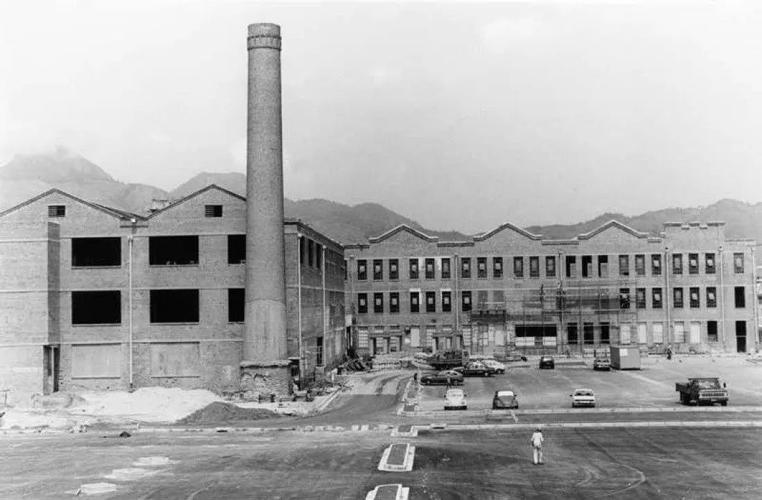 Fábrica industrial com edifícios, chaminé e montanhas ao fundo.