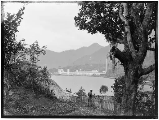 Uma fotografia antiga em preto e branco mostra vegetação, água, construção industrial, árvore desidratada, montanhas e um homem olhando para a paisagem.