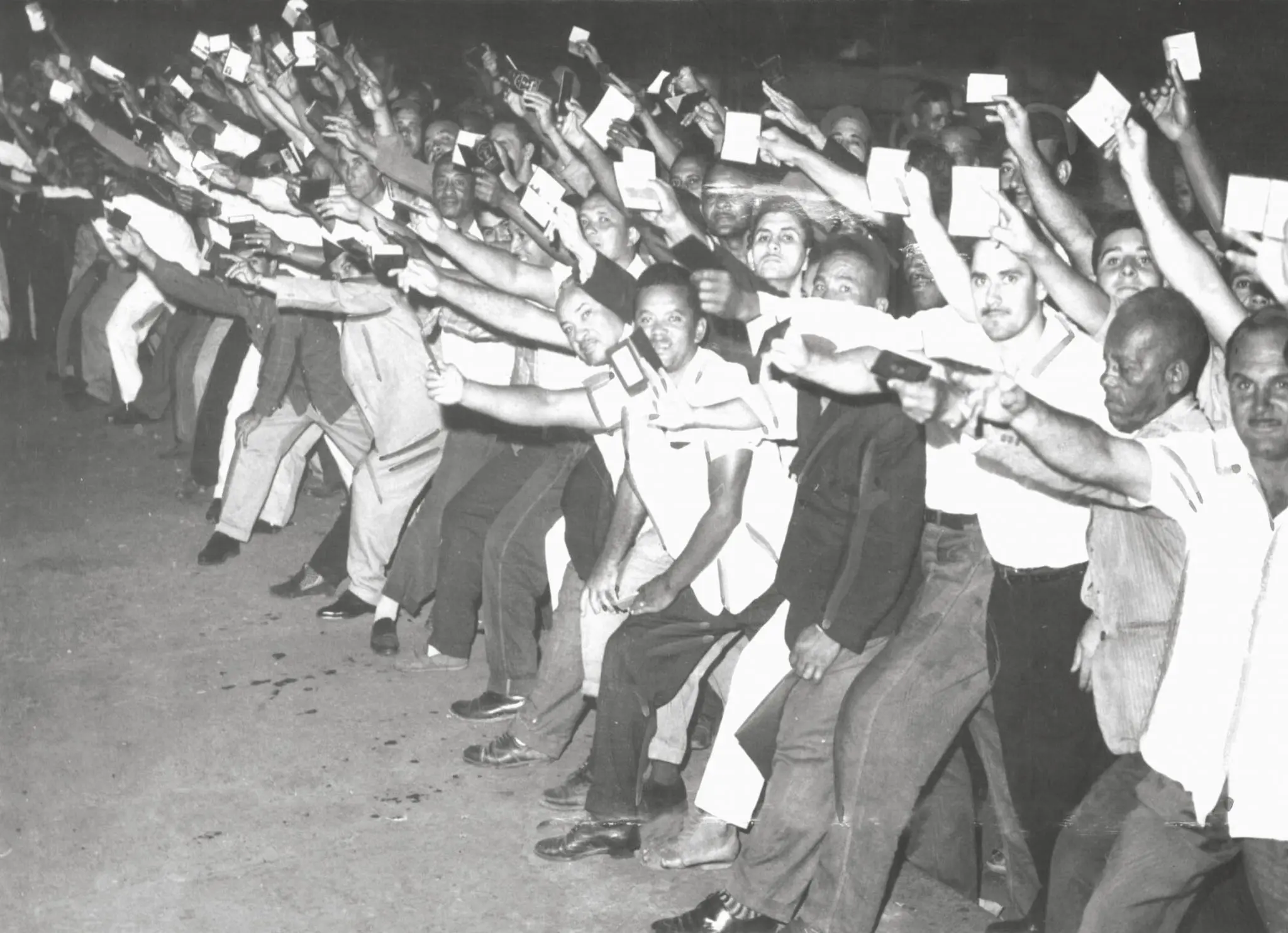 Multidão festiva em movimento, braços levantados, objetos brancos nas mãos.
