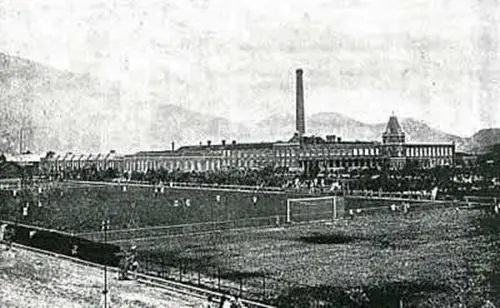 Estádio antigo com arquibancadas, campo de futebol e edifício industrial.