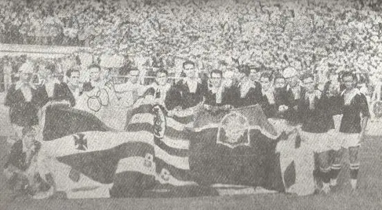 Grupo de pessoas em uniforme esportivo posando em estádio, segurando bandeira.