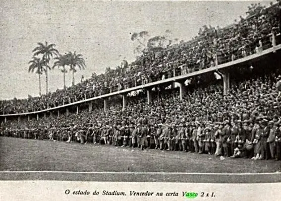 Estádio lotado durante jogo de futebol, com torcedores em tons escuros e palmeiras ao fundo.