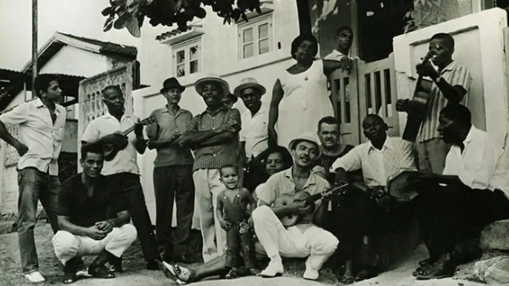 Grupo de pessoas reunidos em frente a uma casa, com músicos tocando instrumentos.