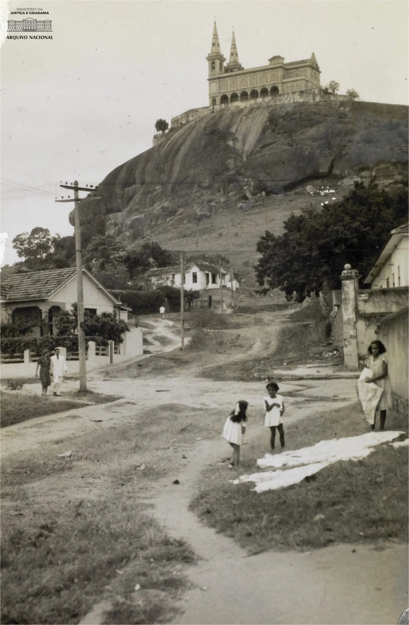Edifício com torres em colina rochosa, casas simples e árvore isolada ao fundo.