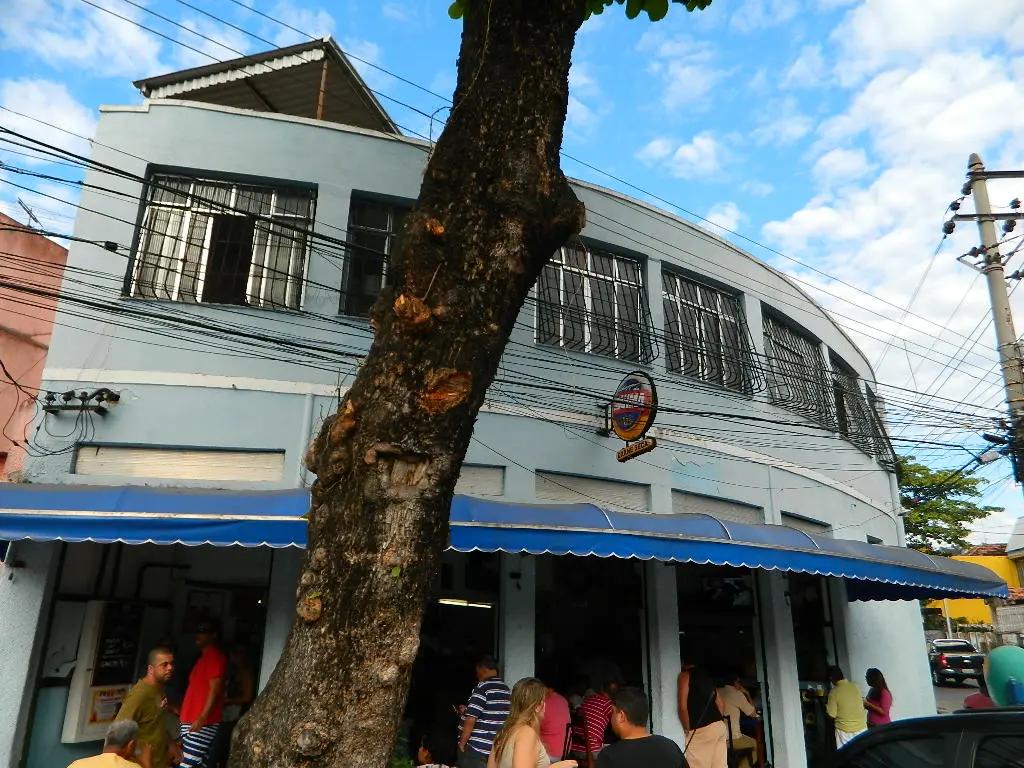 Bar "O Bar Amendoeira" com pessoas na entrada, edifício azul e árvore verde.