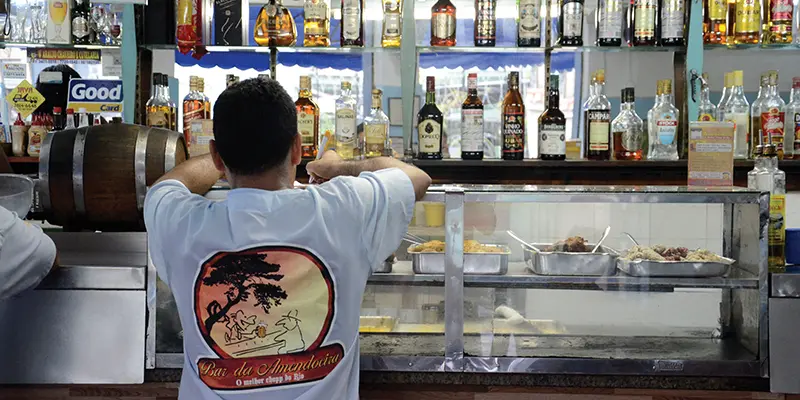 Bar da Amendoeira com vitrine de comida, homem de costas e garrafas de bebidas.