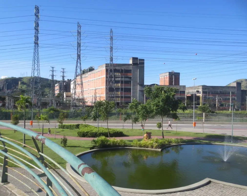 Parque urbano com lagoa, vegetação verde e edifícios industriais ao fundo.