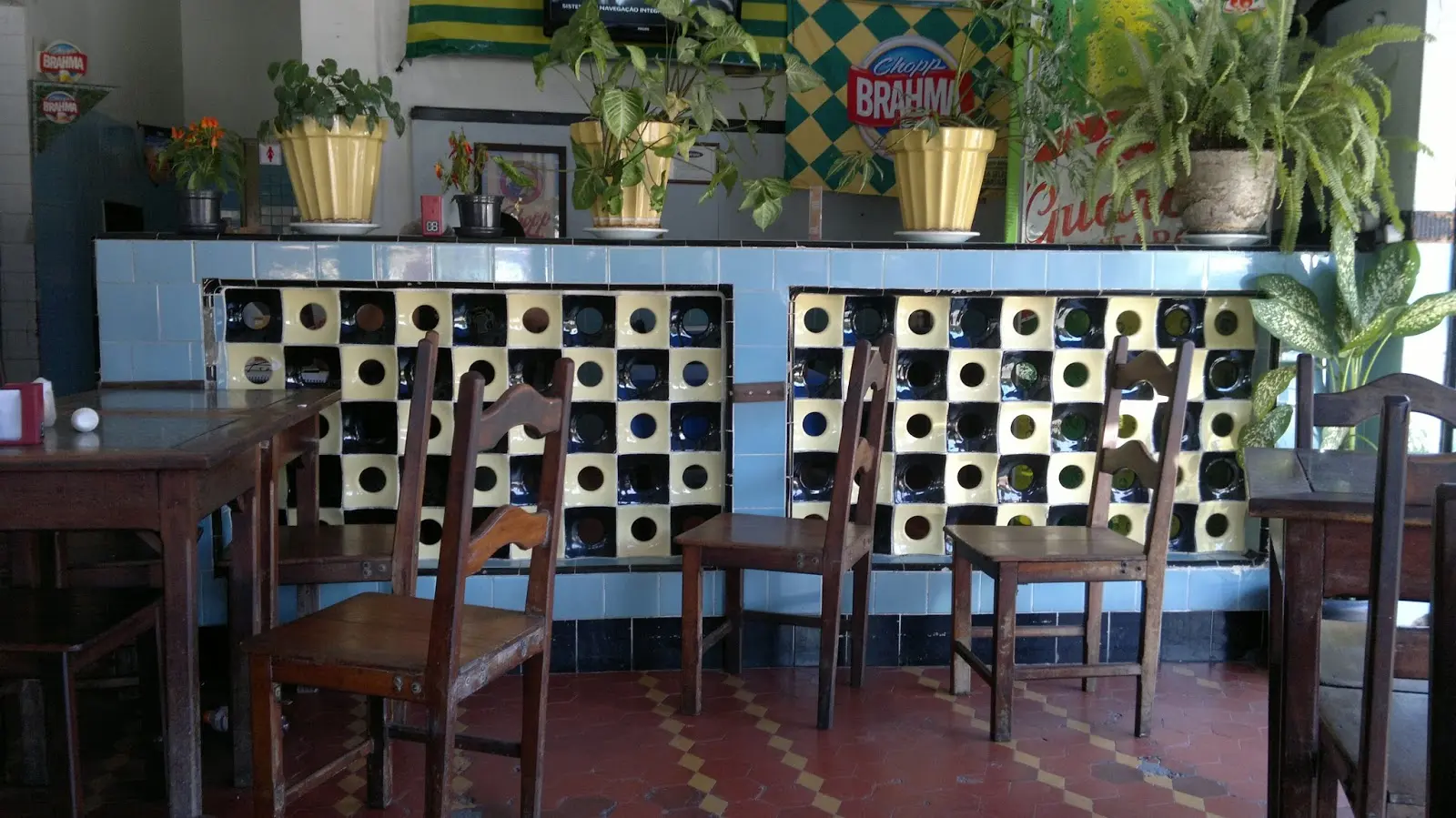 Bar Amendoeira com mesas de madeira, azulejos preto-amarelos e balcão com plantas douradas.
