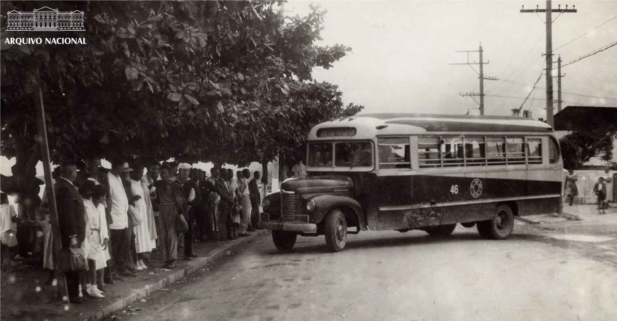 Grupo de pessoas aguardando ônibus na calçada, com ônibus antigo à direita.