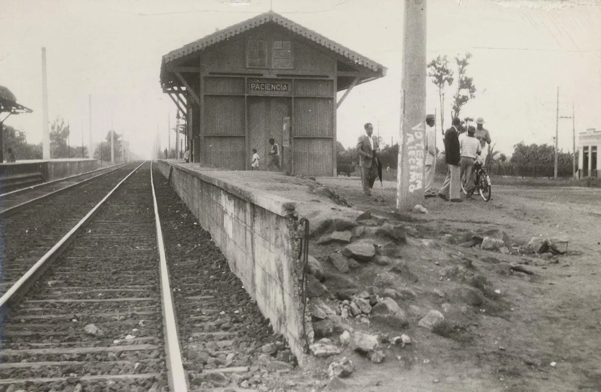 Estação ferroviária com trilhas de trens, muro de concreto, pequena construção e árvores ao fundo.
